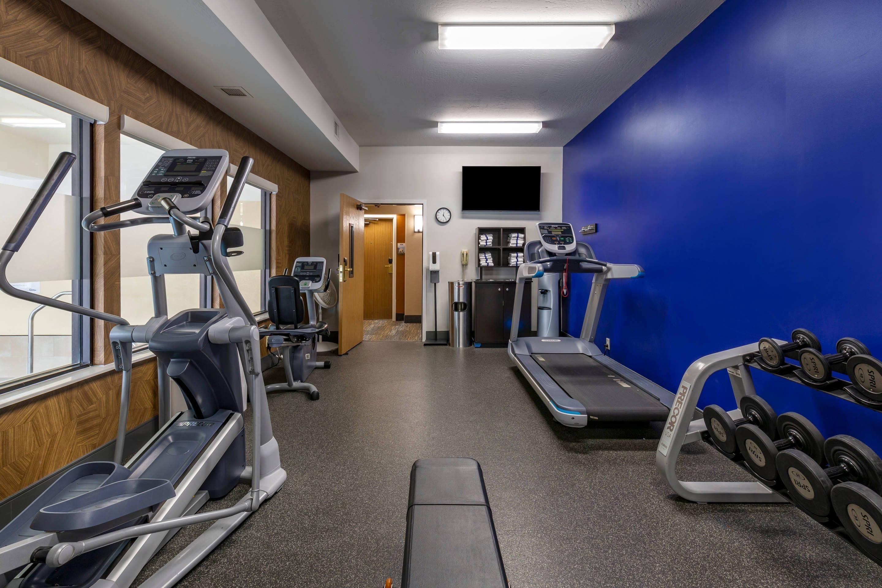 A gym with treadmills, elliptical machines, and a weight bench with dumbbells.