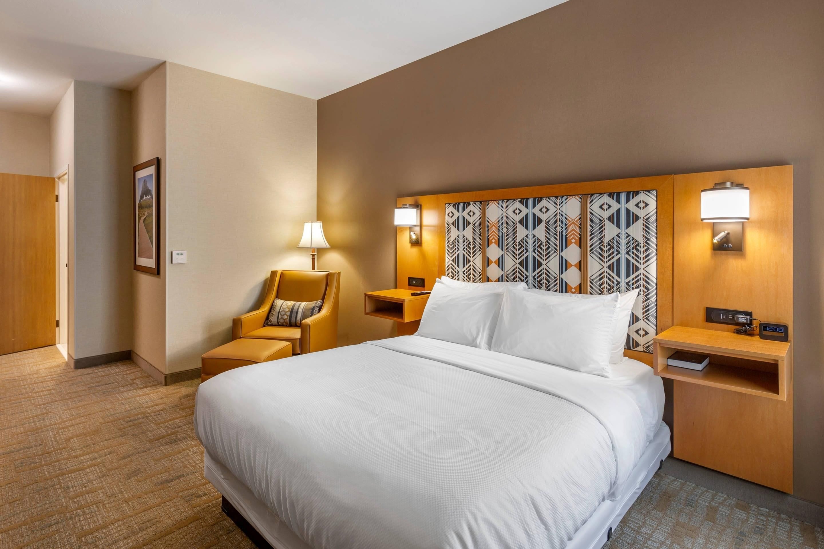 Quarto de hotel com uma cama grande, uma cadeira, uma mesa de cabeceira e uma luminária fixada na parede.