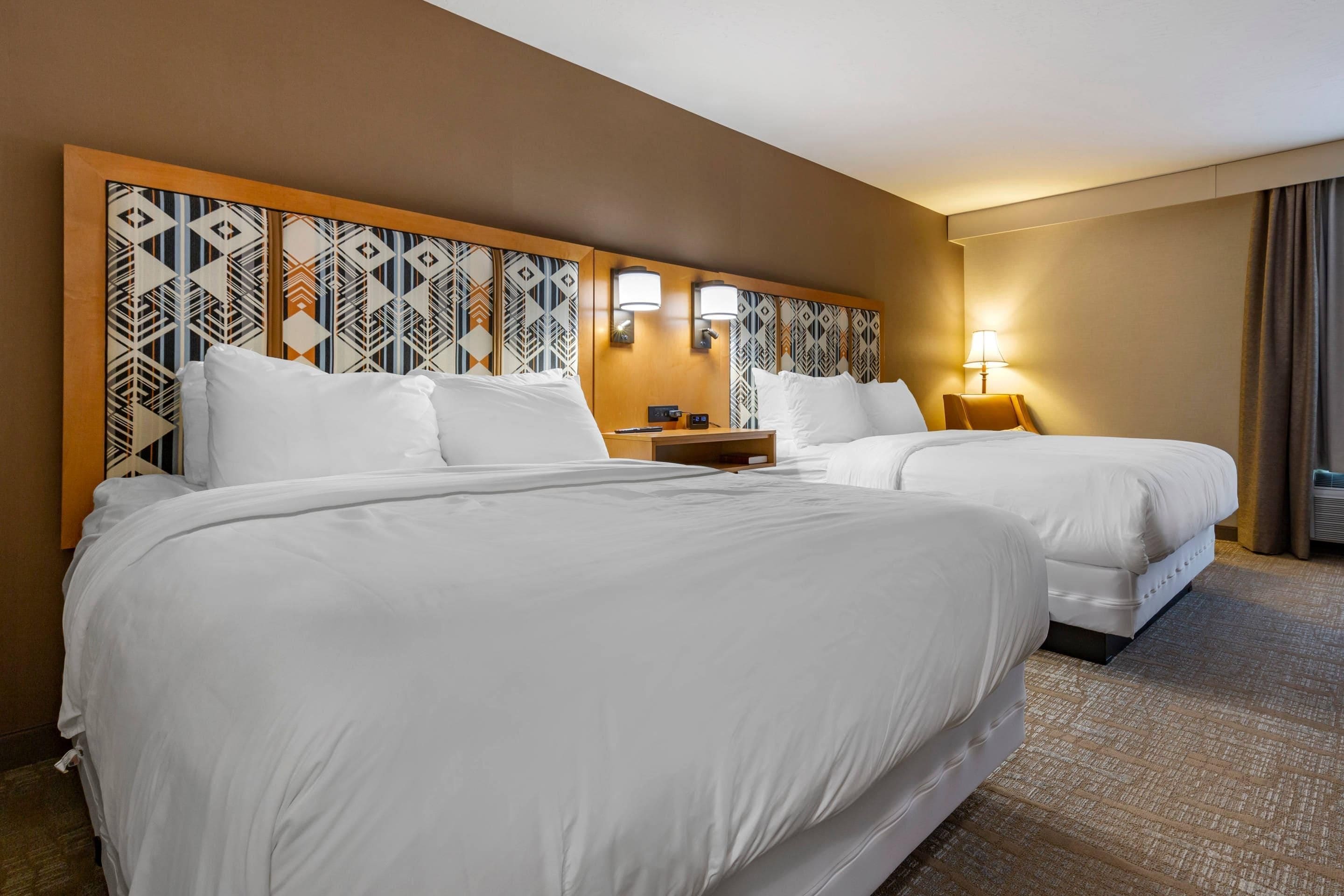 A hotel room with two beds, a headboard with a geometric pattern, bedside lamps, and a carpeted floor.
