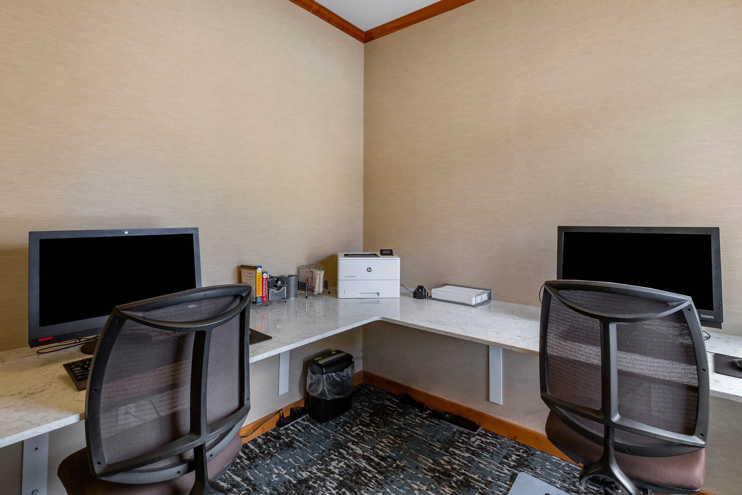 Ein Hotelzimmer mit zwei Schreibtischen, jeweils ausgestattet mit einem Computermonitor, einer Tastatur und einem Stuhl. Auf dem Schreibtisch steht ein Drucker, und ein Papierkorb ist sichtbar. Der Raum hat einen Teppichboden und eine Holzbodenverkleidung.