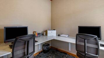 Ein Hotelzimmer mit zwei Schreibtischen, jeweils ausgestattet mit einem Computermonitor, einer Tastatur und einem Stuhl. Auf dem Schreibtisch steht ein Drucker, und ein Papierkorb ist sichtbar. Der Raum hat einen Teppichboden und eine Holzbodenverkleidung.