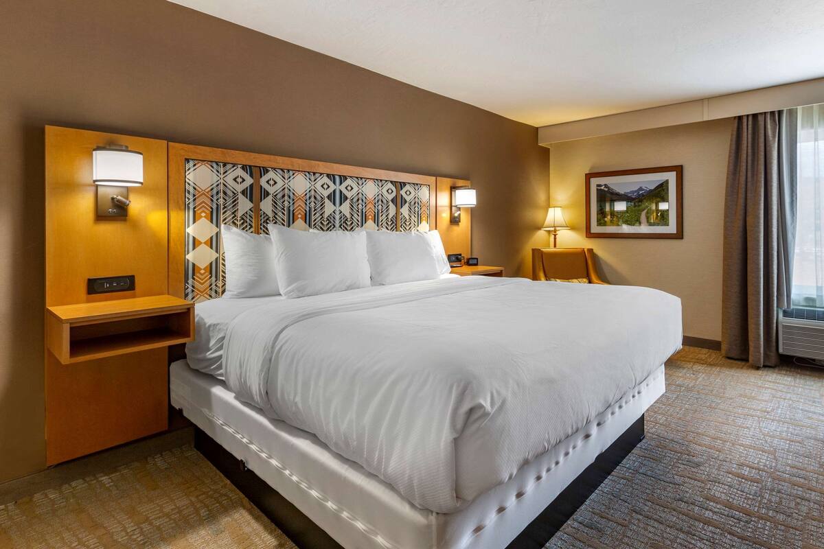 A hotel room with a large bed, wooden headboard, two bedside tables, a nightstand, a lamp, a picture on the wall, and a window with curtains.