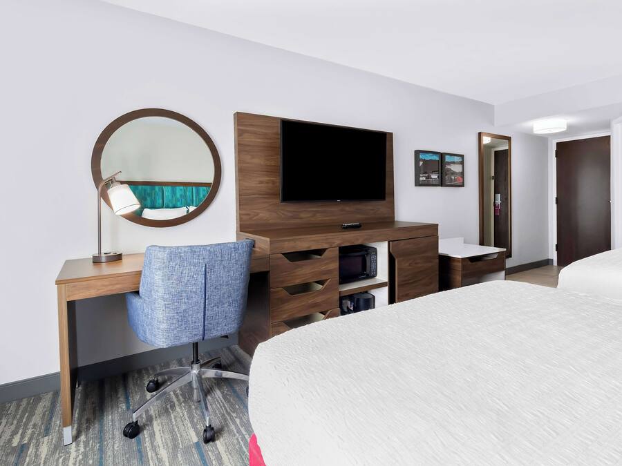 Una habitación de hotel con cama, escritorio con silla, televisor de pantalla plana, espejo, microondas y puerta.