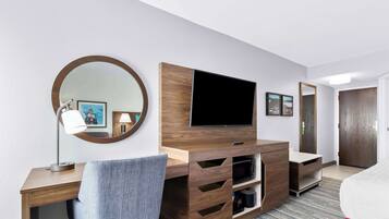 Habitación de hotel con un escritorio de madera, un televisor de pantalla plana, un espejo redondo y una cama con una almohada roja decorativa.