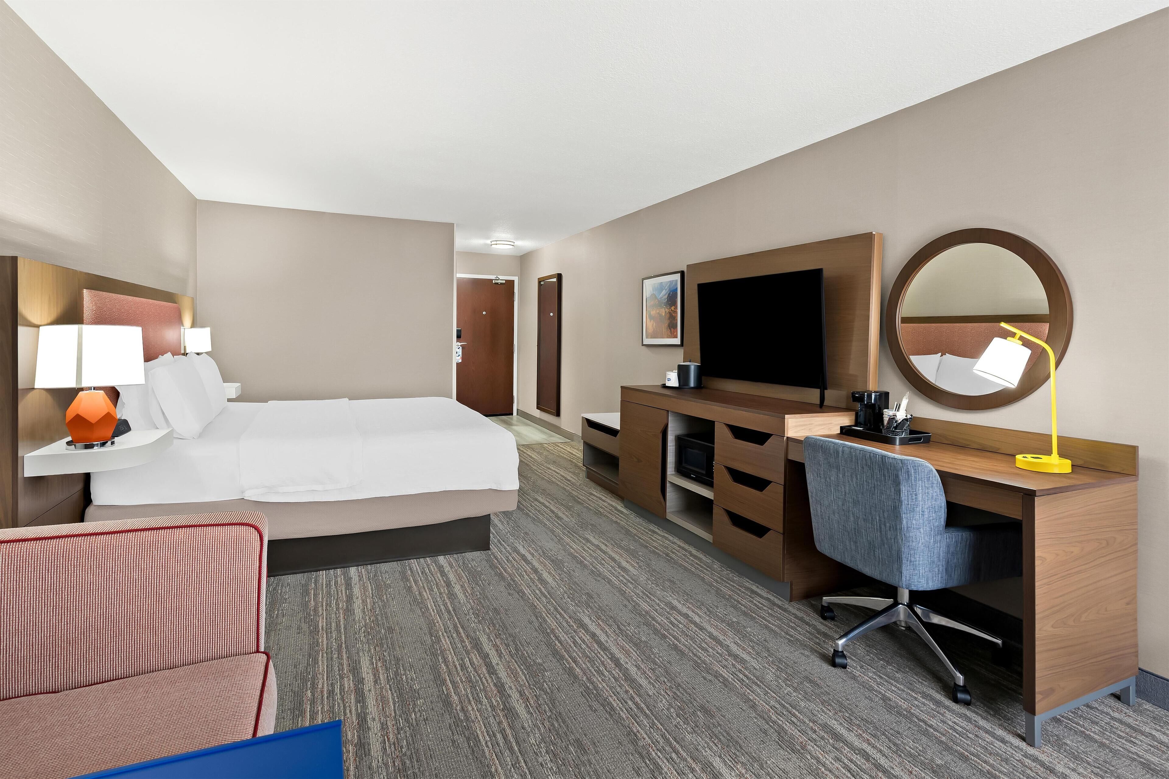 A hotel room with a large bed, a desk with a chair, a TV, a desk lamp, and a mirror.