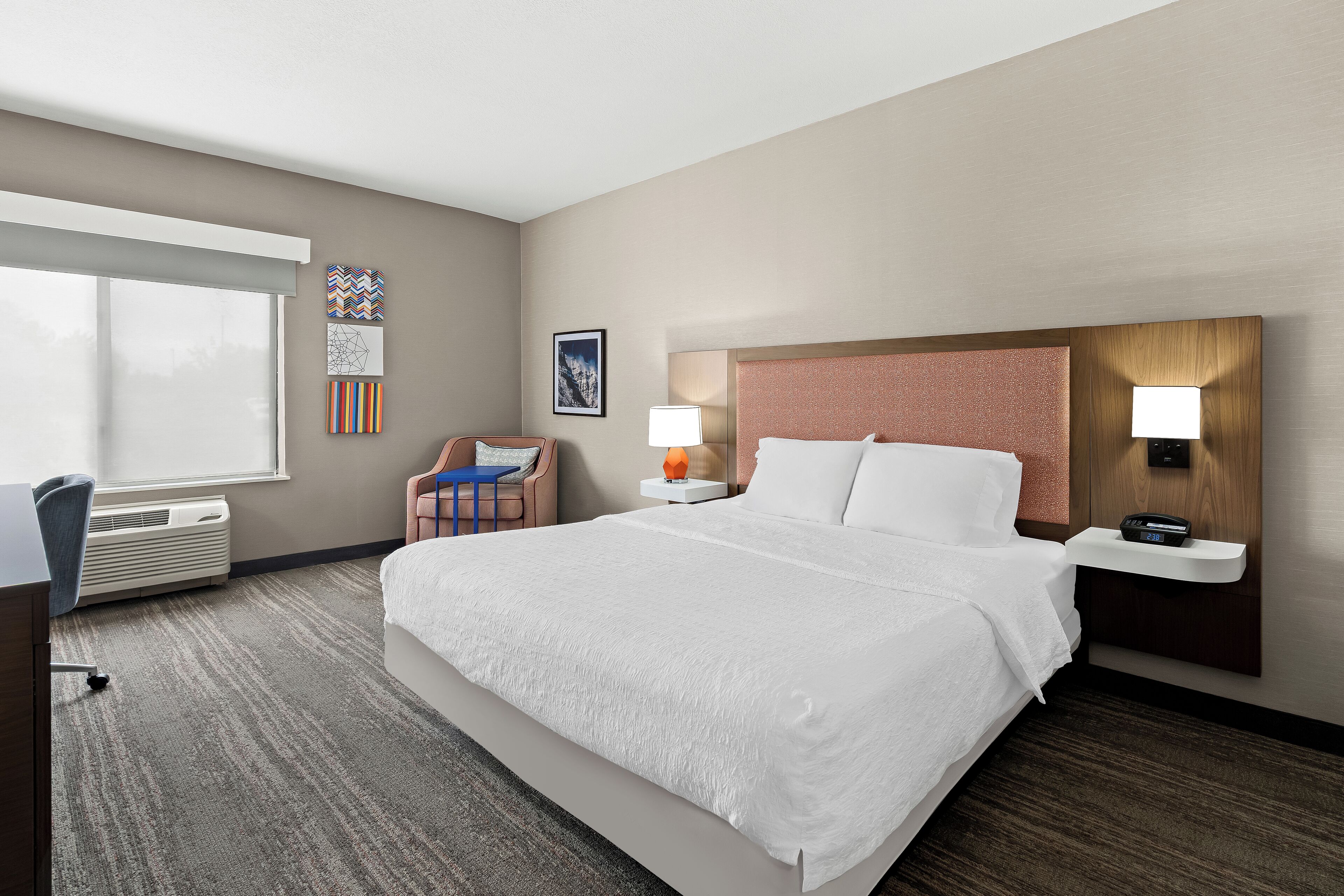 A hotel room with a large bed, a desk, a chair, a window, and a wall with artwork.
