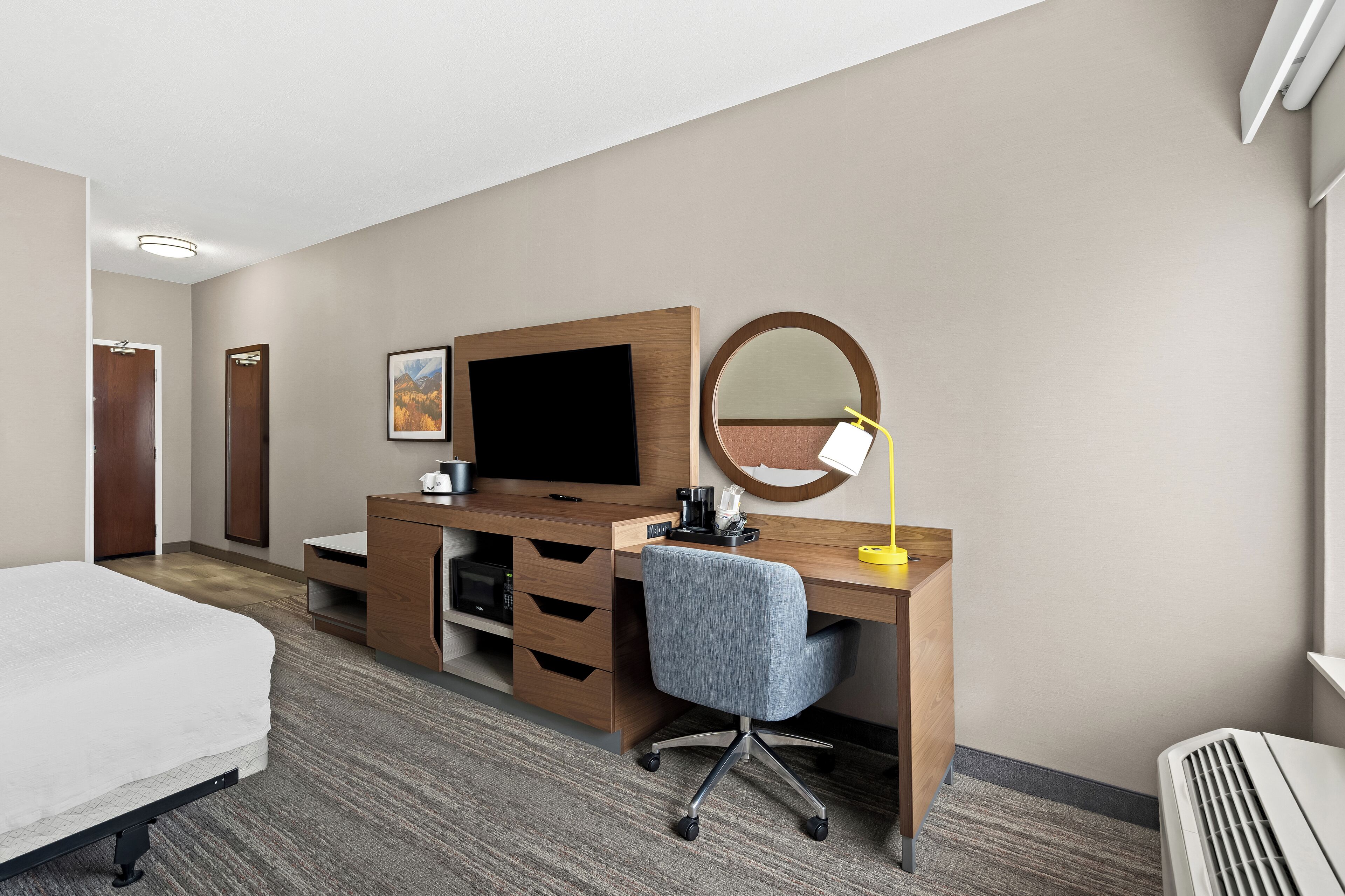 Een hotelkamer met een bed, een bureau met een computer, een bureaulamp, een spiegel en een televisie.