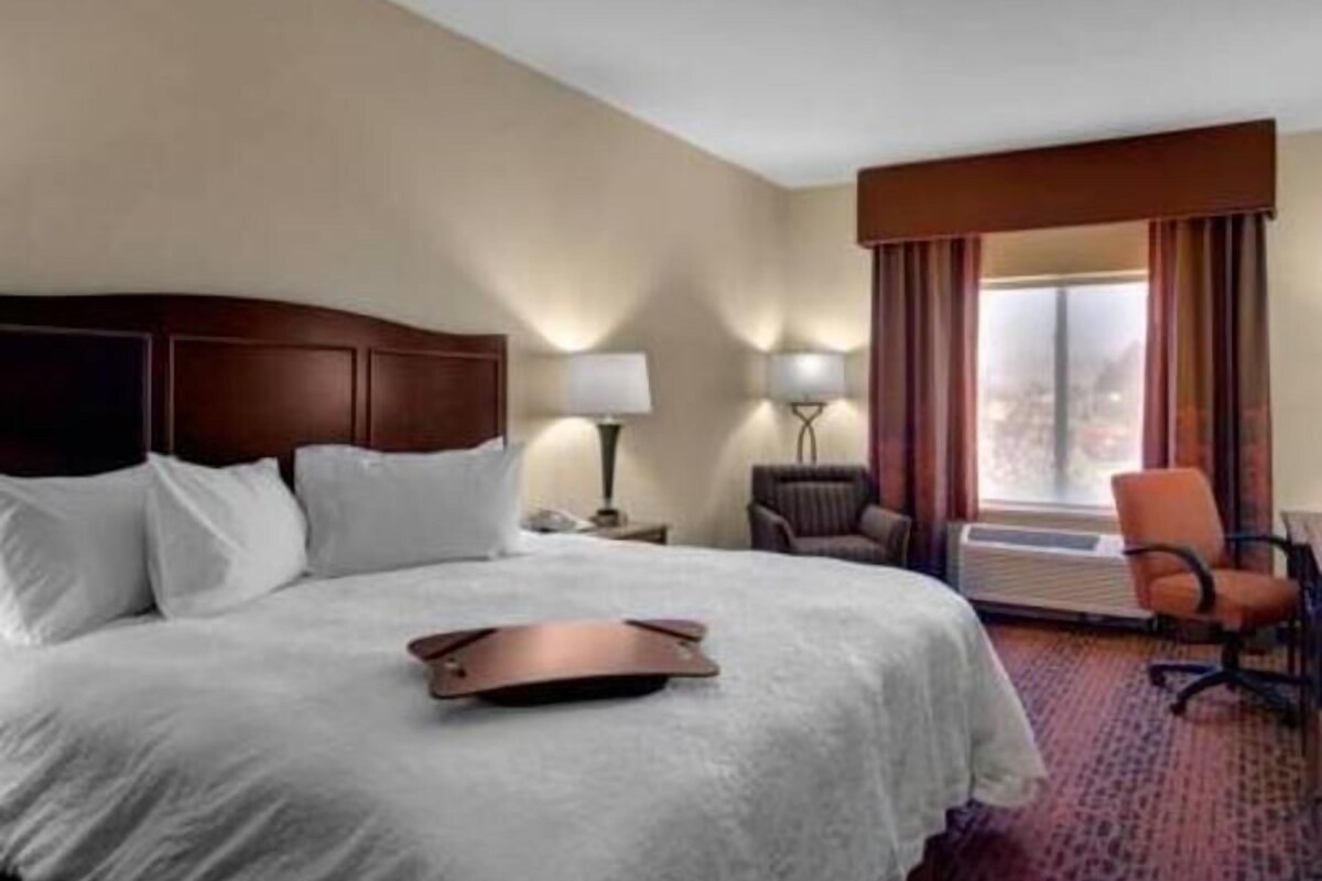 Habitació d'hotel amb un llit, escriptori amb làmpada, cadira i finestra amb cortines.