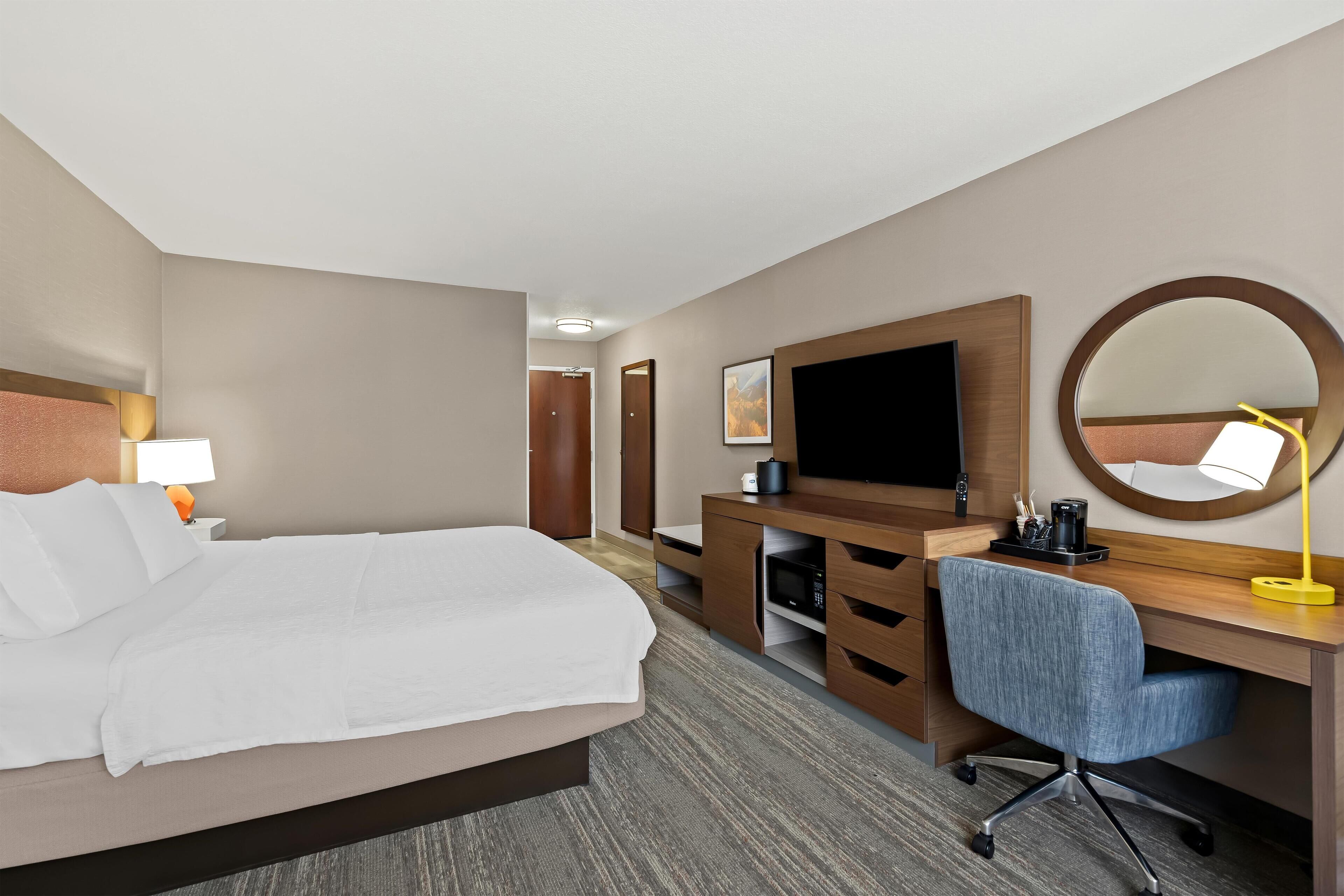 A hotel room with a bed, a desk, a chair, a television, a mirror, and a lamp.