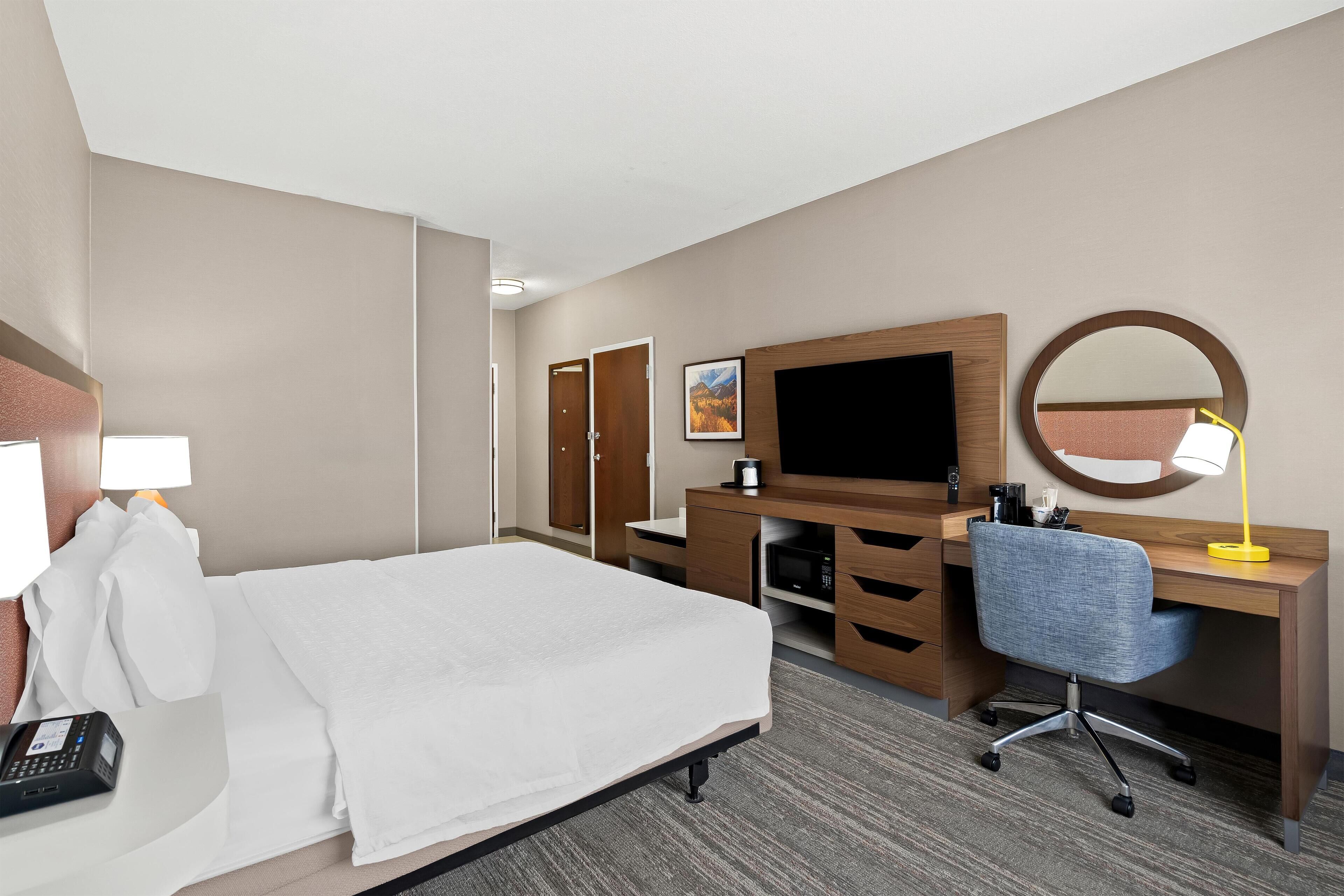 A hotel room with a bed, a desk with a computer, a TV, a mirror, and a lamp.