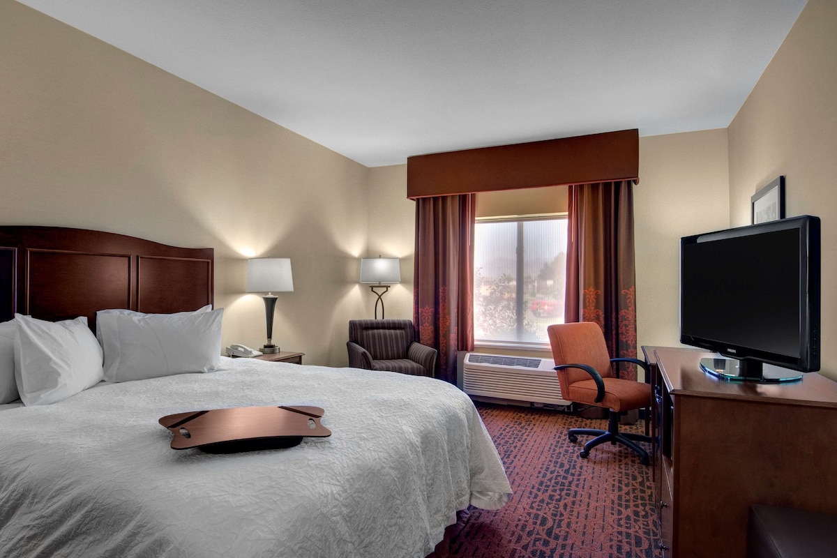 Une chambre d’hôtel avec un lit, un bureau avec une télévision, une chaise, une lampe et une fenêtre avec des rideaux.