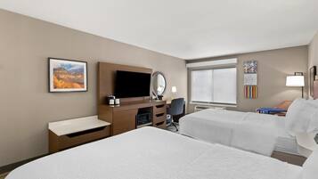 Quarto de hotel com duas camas, uma escrivaninha com televisão de tela plana, um espelho e uma janela com persianas.
