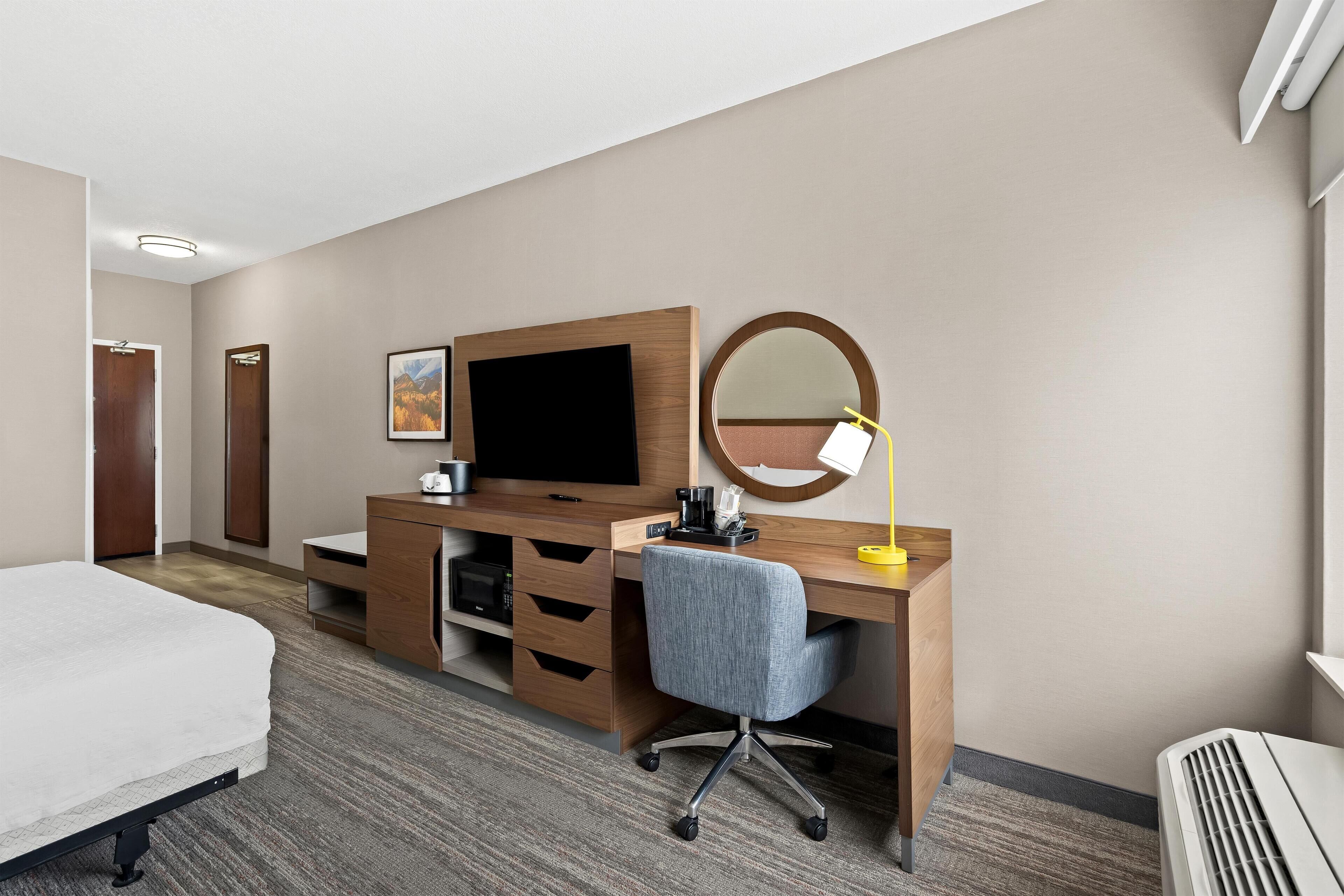 Quarto de hotel com cama, escrivaninha com computador, cadeira, abajur, espelho e televisão.