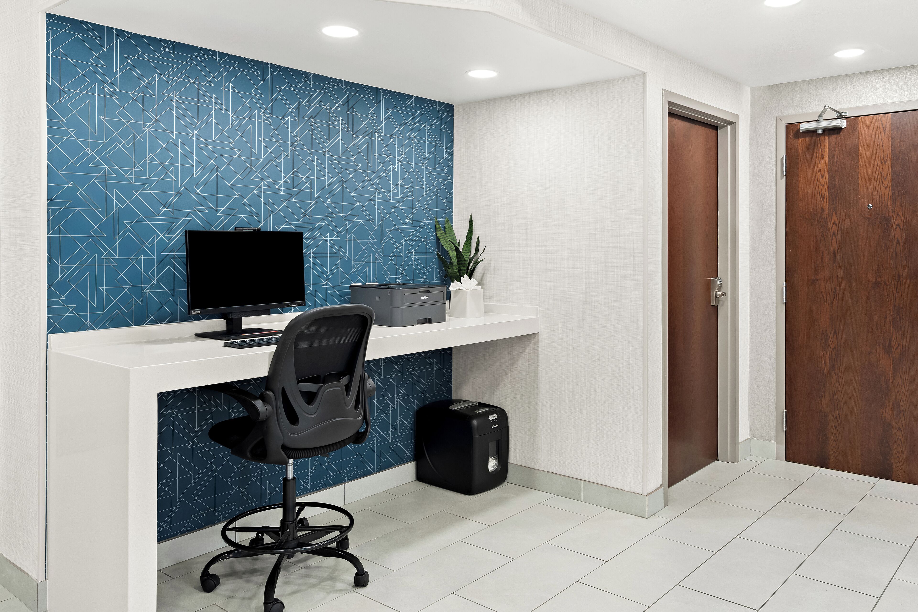 A modern office space with a blue geometric wall, a white desk, a black office chair, a black trash bin, and a wooden door.