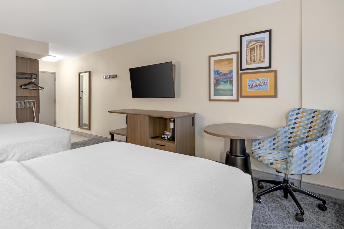 A hotel room with two beds, a desk, a chair, a TV, and framed pictures on the wall.