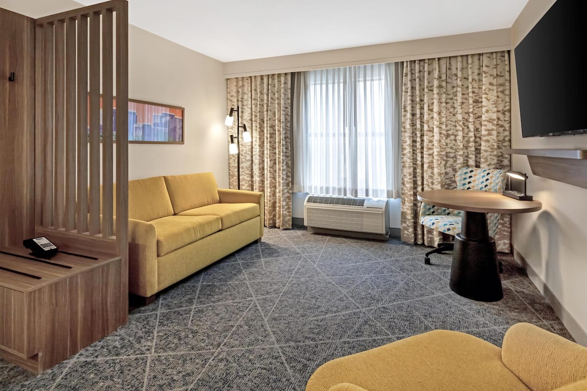 A hotel room with a sofa, a small round table with a lamp, a television mounted on the wall, and a large window with curtains.