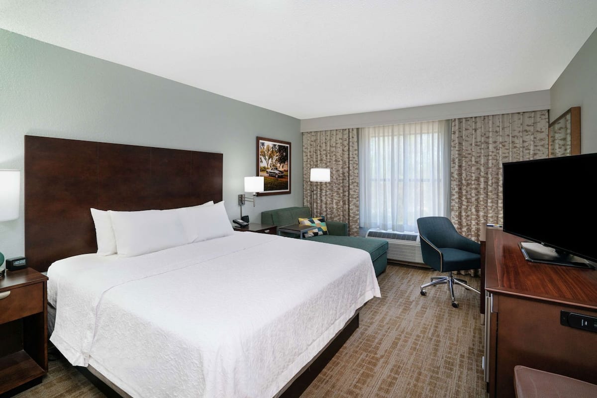 A hotel room with a large bed, a desk with a TV, a chair, a desk lamp, a nightstand, and a window with curtains.
