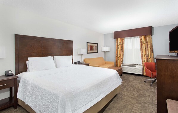 A hotel room with a large bed, a desk with a lamp, a chair, a television, and a window with curtains.