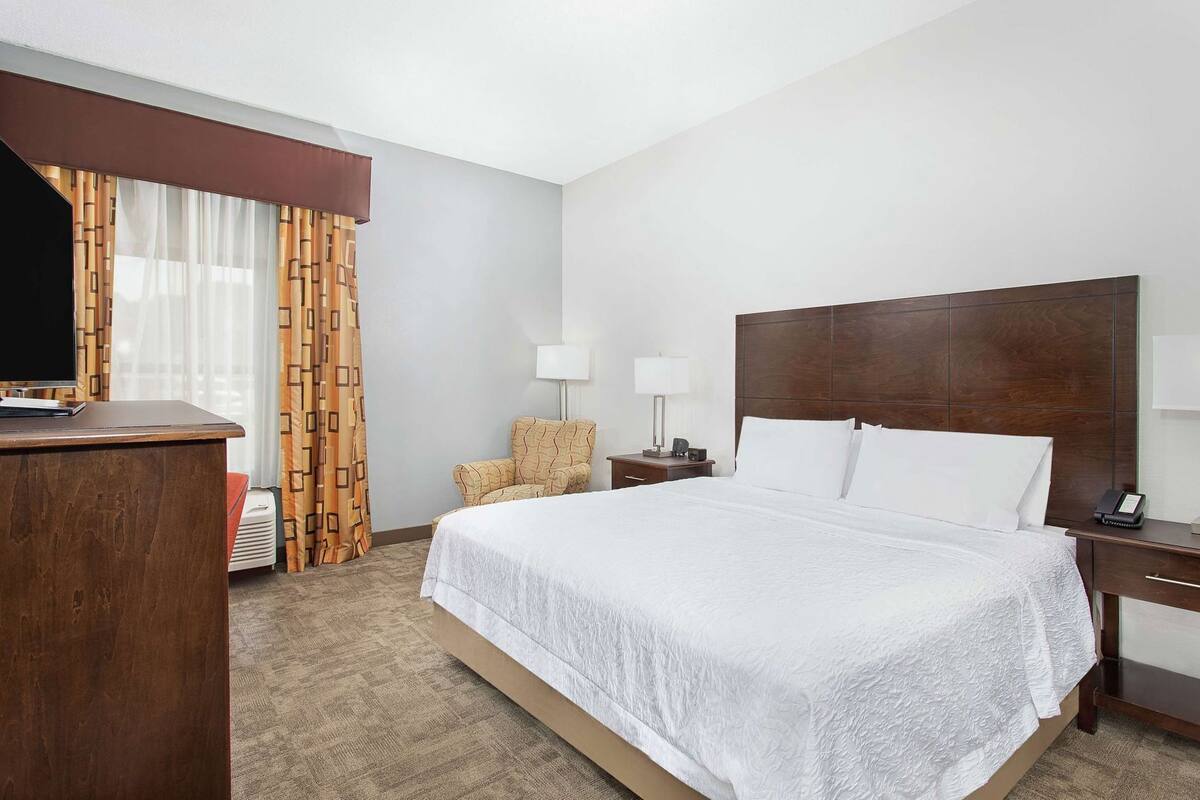 Una habitación de hotel con una cama grande, un escritorio con televisor, una silla, una mesita de noche con lámpara y una ventana con cortinas.
