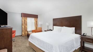 A hotel room with a large bed, a desk with a TV, a chair, a nightstand with a lamp, and a window with curtains.