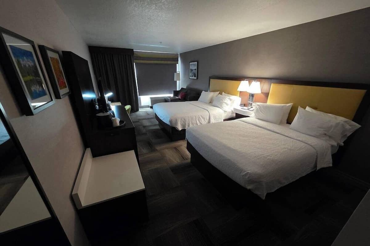 A hotel room with two beds, a desk, a chair, a lamp, and a picture on the wall.