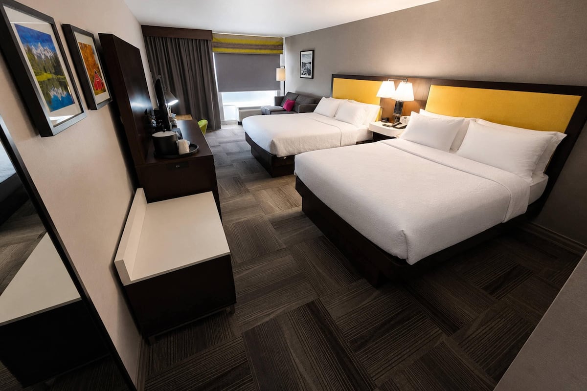 A hotel room with two beds, a desk, a chair, a lamp, and artwork on the walls.