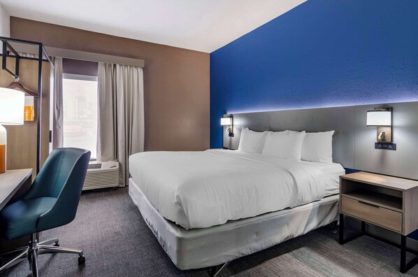 A hotel room with a large bed, a desk with a chair, a nightstand, and a window with curtains.
