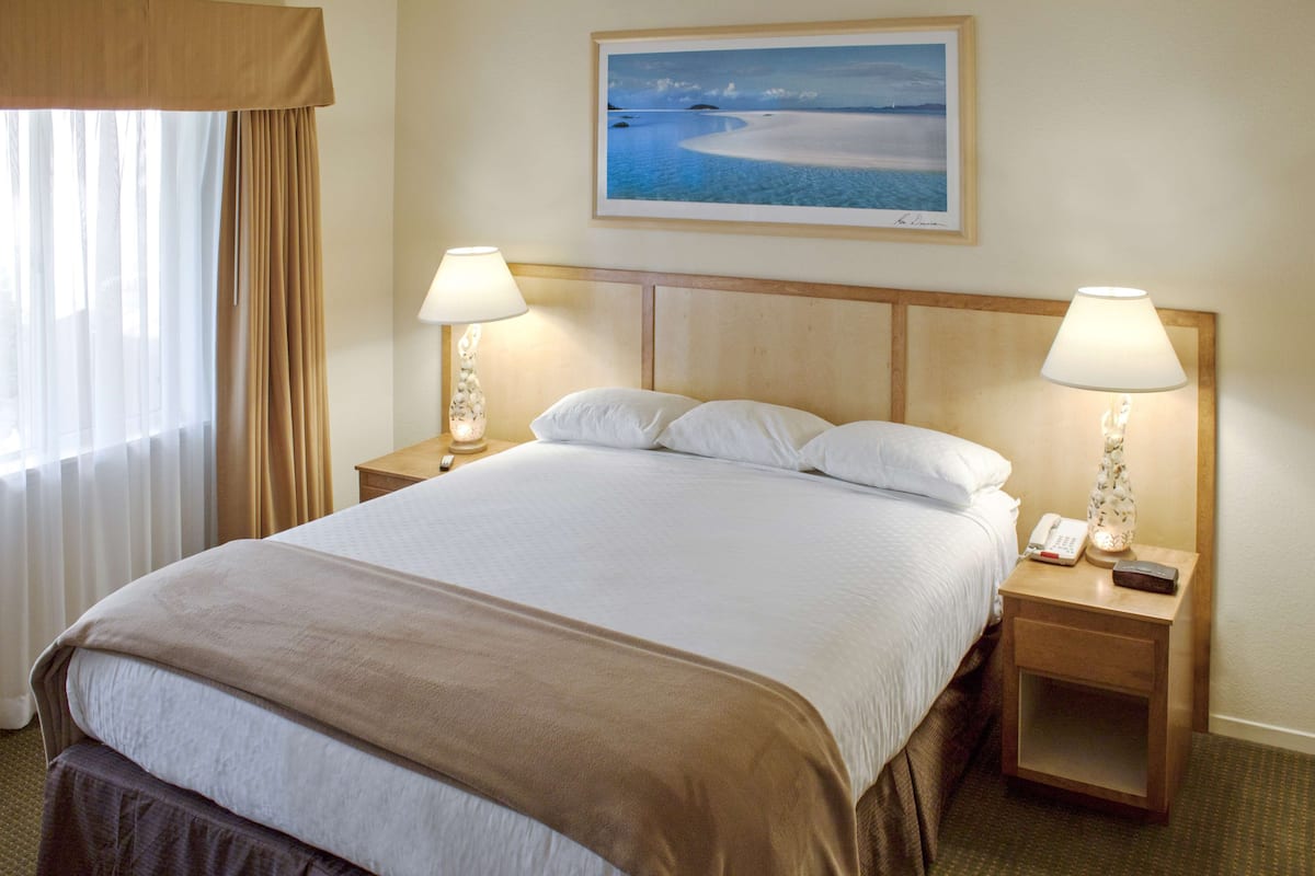 A hotel room with a large bed, two bedside tables with lamps, a painting of a beach scene, and a window with curtains.