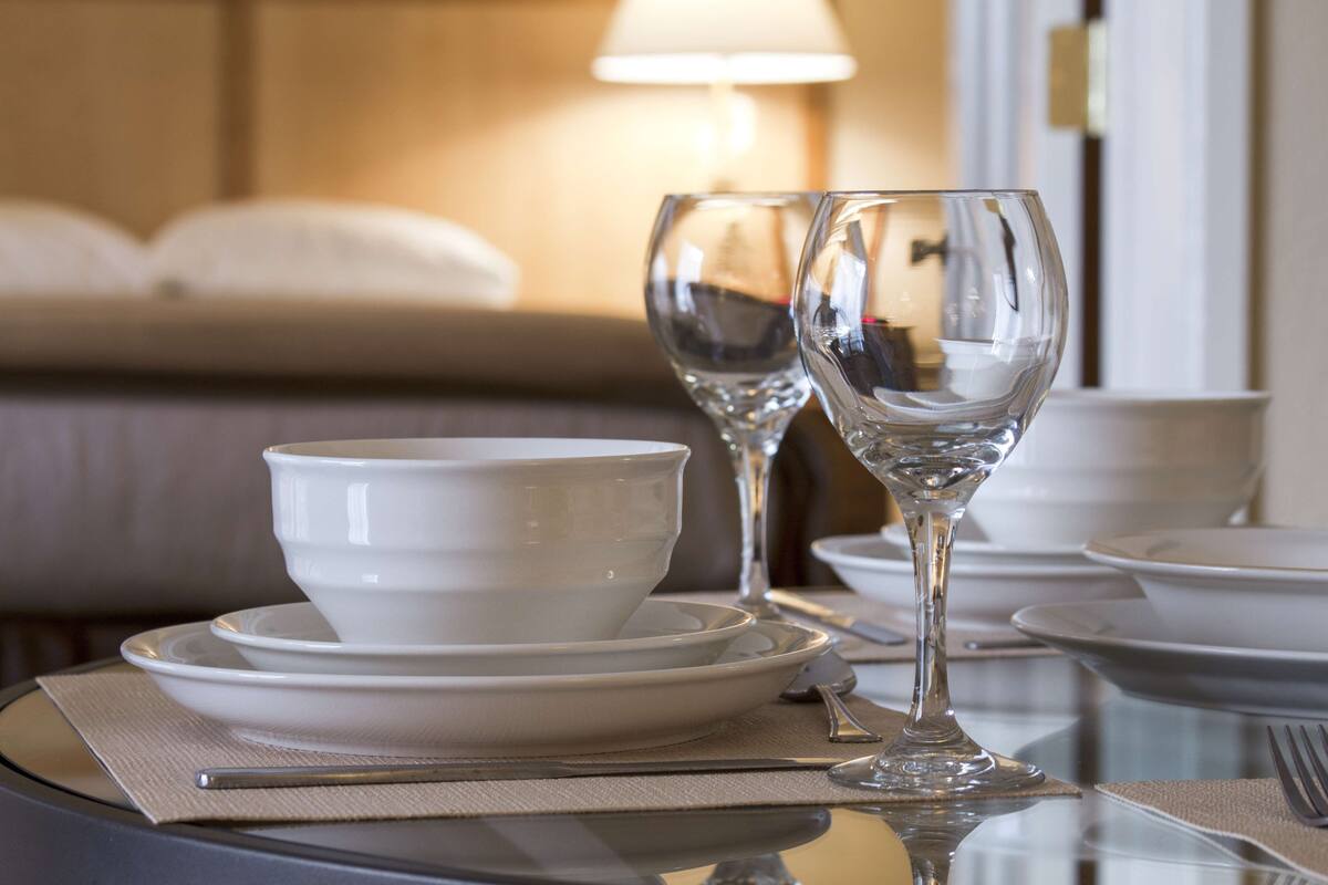 A dining table set with white plates, bowls, and wine glasses, with a lamp in the background.