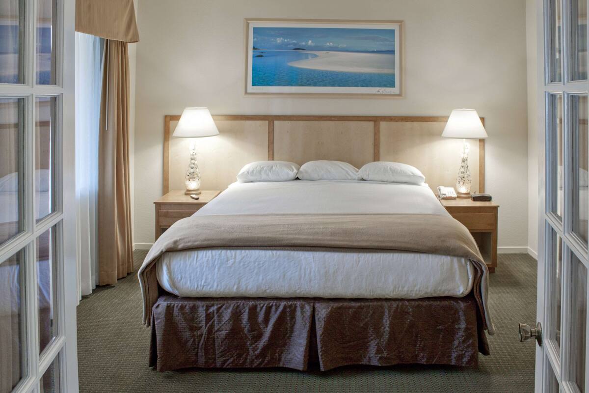 A neatly made bed with a headboard, two bedside lamps, and a framed picture above it.