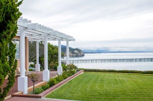 A white pergola with columns, a well-maintained lawn, and a view of a body of water with a pier in the distance.