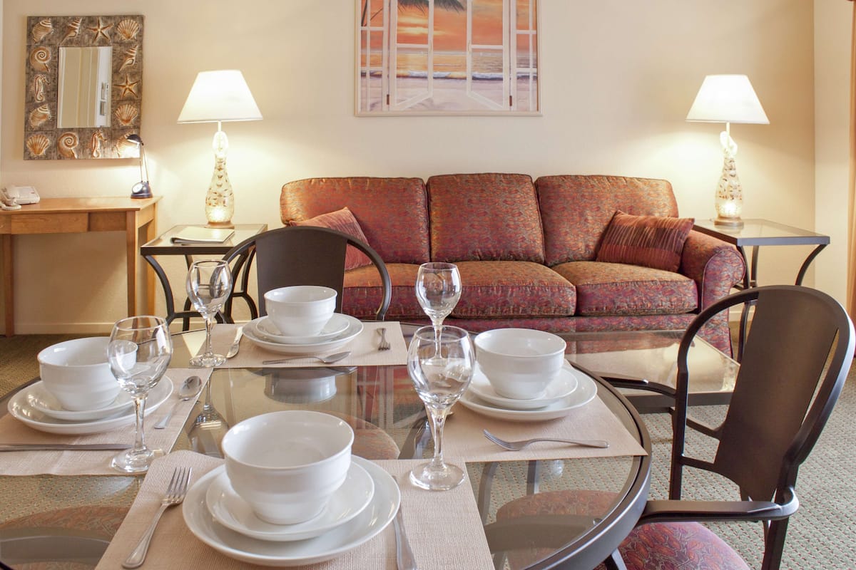 A dining area with a round glass table set for four, surrounded by a patterned sofa and two lamps.