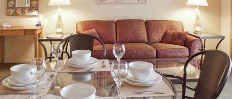 Un coin repas avec une table ronde en verre, dressée pour quatre personnes, entourée d’un canapé à motifs et de deux lampes.