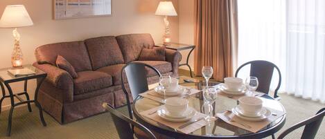 A hotel room with a dining table set for four, a sofa, two lamps, and a large window with curtains.