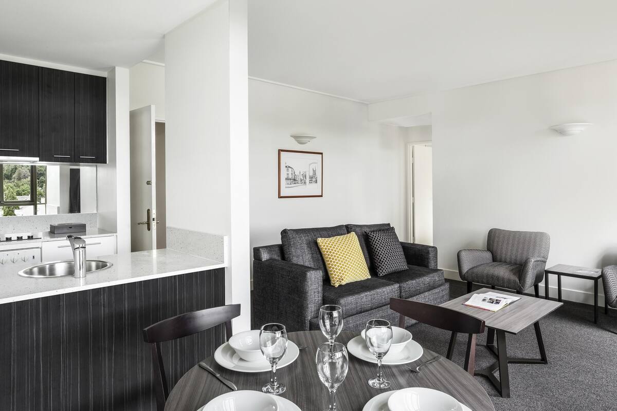 Een moderne keuken met donkere kastjes, een roestvrijstalen spoelbak en een eethoek met een ronde tafel voor twee personen.