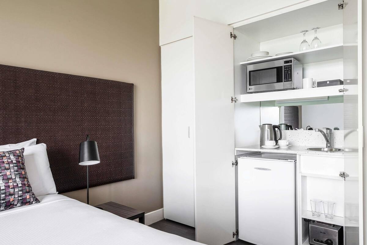 Een compacte hotelkamer met een bed, bedlampje, een aan de muur bevestigde televisie, een kitchenette met een magnetron, waterkoker en een kleine koelkast.