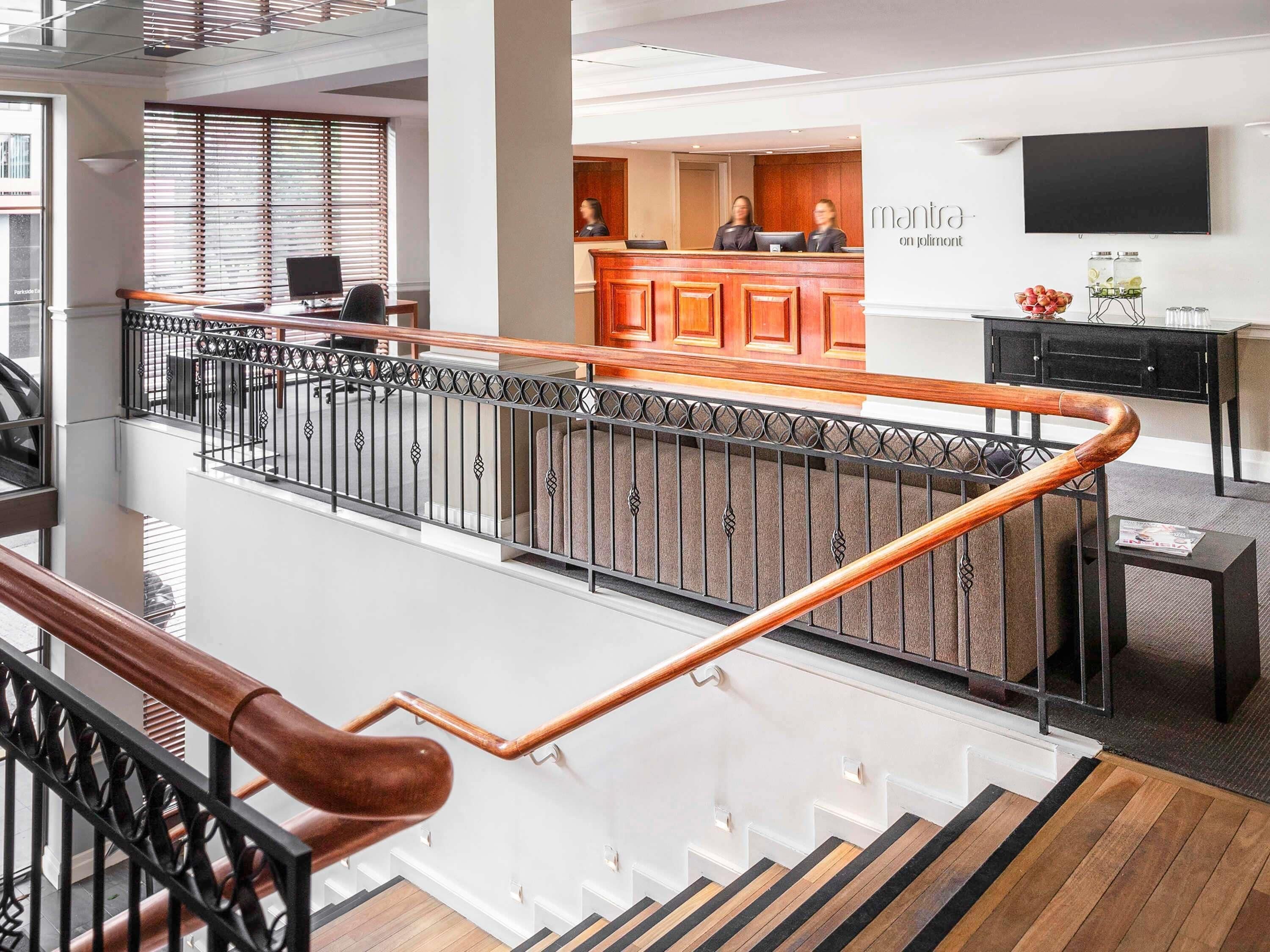A modern hotel lobby with a reception desk, a staircase with wooden handrails, and a flat-screen TV on the wall.