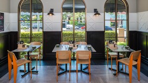 A restaurant with a modern interior, featuring a central dining table surrounded by chairs, large arched windows, and a view of the street outside. - Mantra on Jolimont Melbourne (East Melbourne)