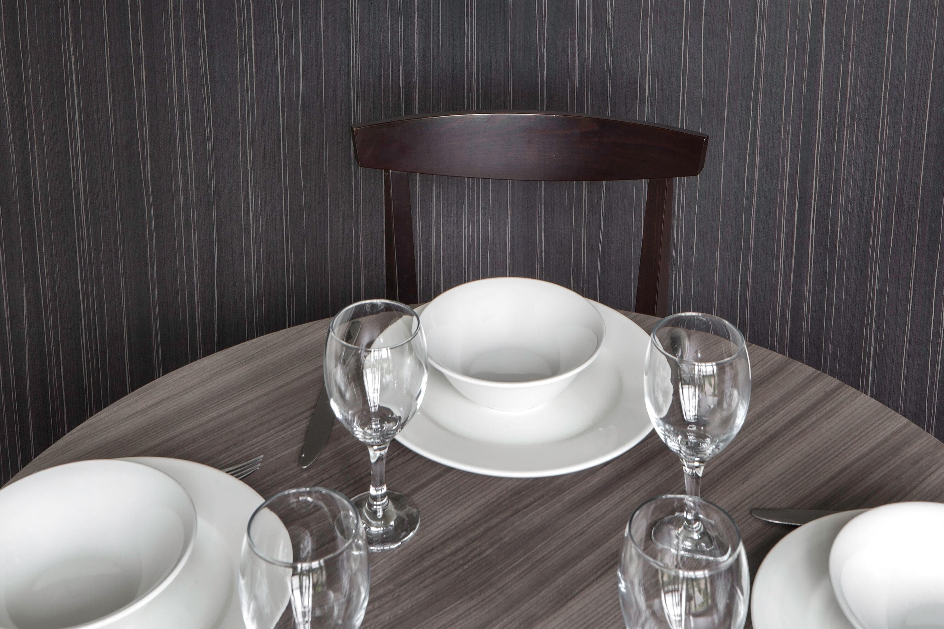 Une table à manger dressée avec de l’assiette blanche, des bols et des verres à vin, et une chaise en bois foncé en arrière-plan.