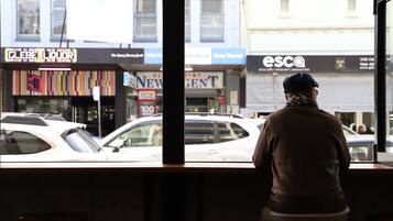 A person seated at a table inside a building, looking out through a window at a street with various shops and cars.
