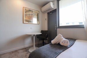 A hotel room with a bed, a small round table, a chair, an air conditioning unit, and a window with curtains.