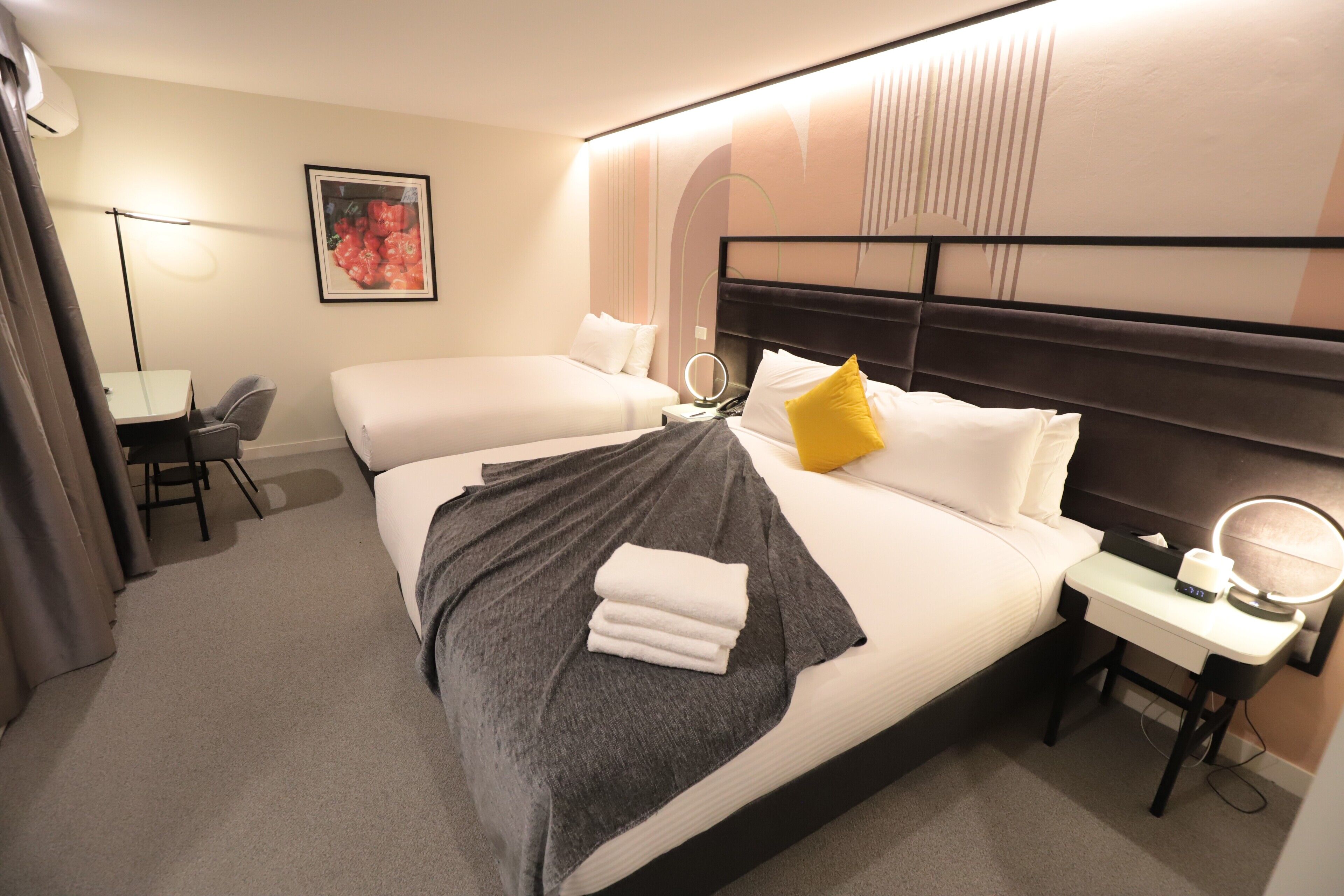 A hotel room with two beds, a desk, a chair, a bedside table with a lamp, and a wall-mounted mirror.