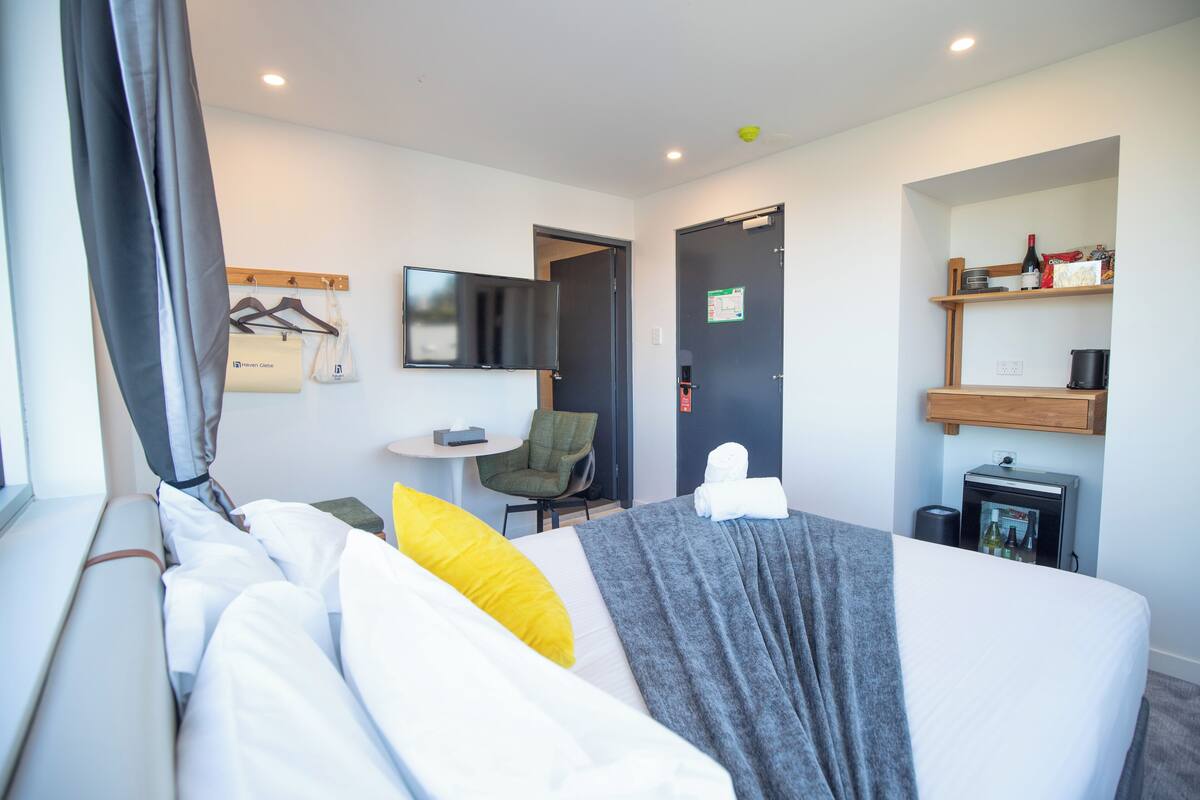 A hotel room with a bed, a chair, a small table, a TV, and a minibar.