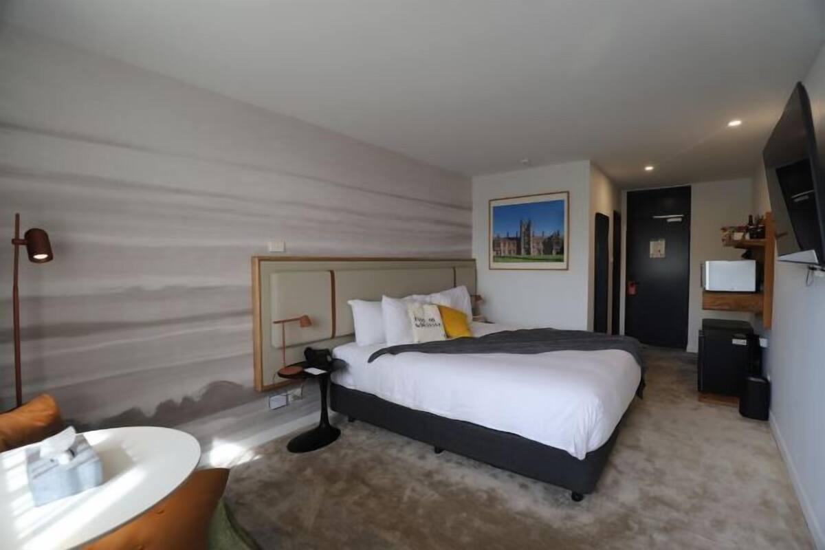 A modern hotel room with a large bed, a desk, a chair, a small table, a lamp, and a painting on the wall.