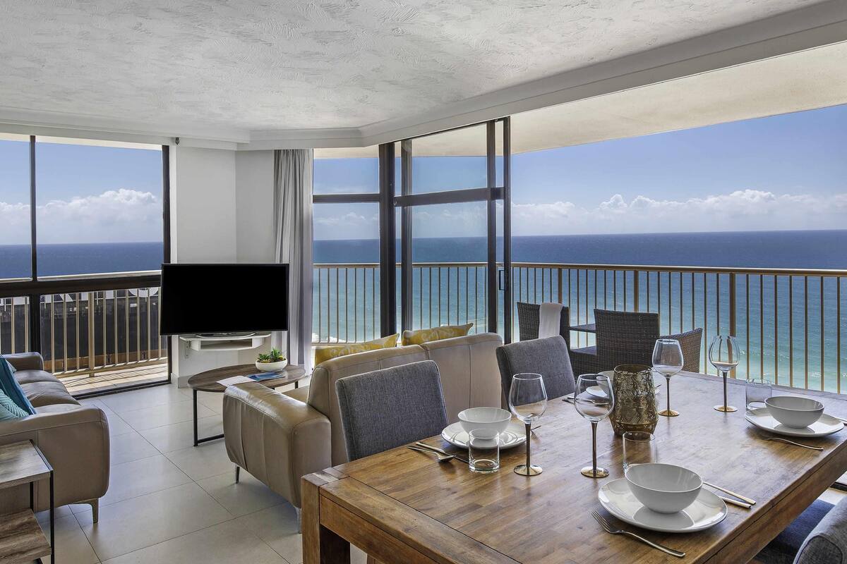 A modern living room with a large TV, comfortable seating, a dining area with table set for two, and a balcony with a view of the ocean.