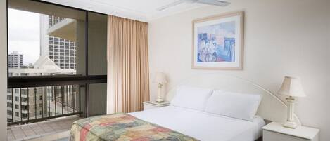 A hotel room with a large bed, a ceiling fan, a window with a view of buildings, a framed picture on the wall, and two bedside lamps.
