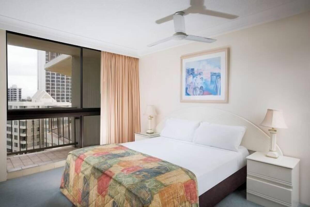 A hotel room with a bed, a ceiling fan, a painting, a window with curtains, and a view of buildings.