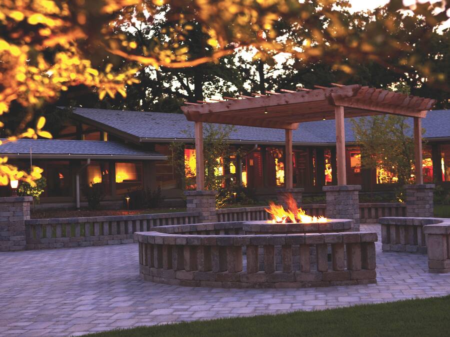 Un espace aménagé autour d’un foyer, avec une pergola, des bancs en pierre et un chemin en briques.