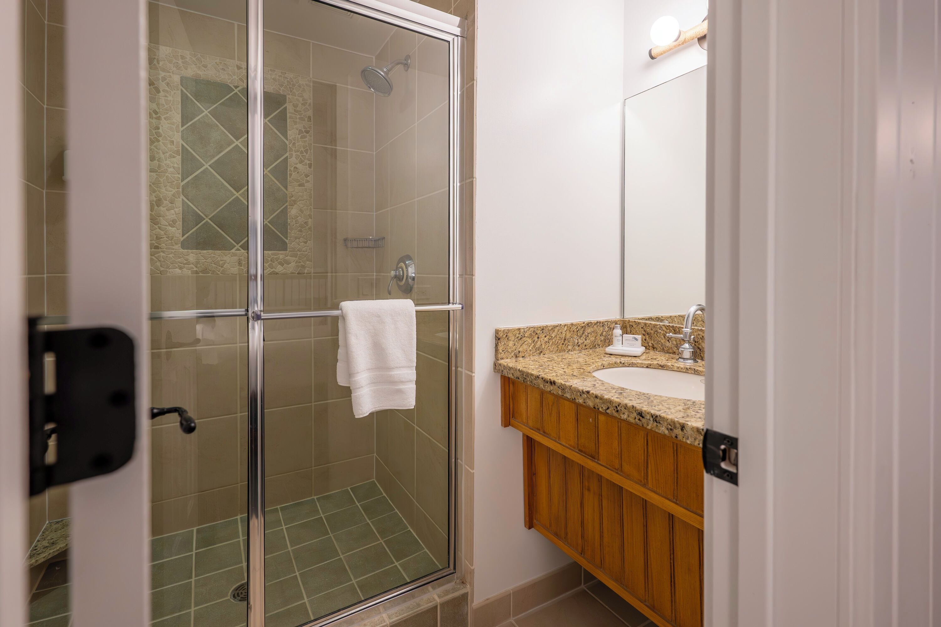 A bathroom with a glass shower enclosure, a sink with a granite countertop, and wooden cabinets.
