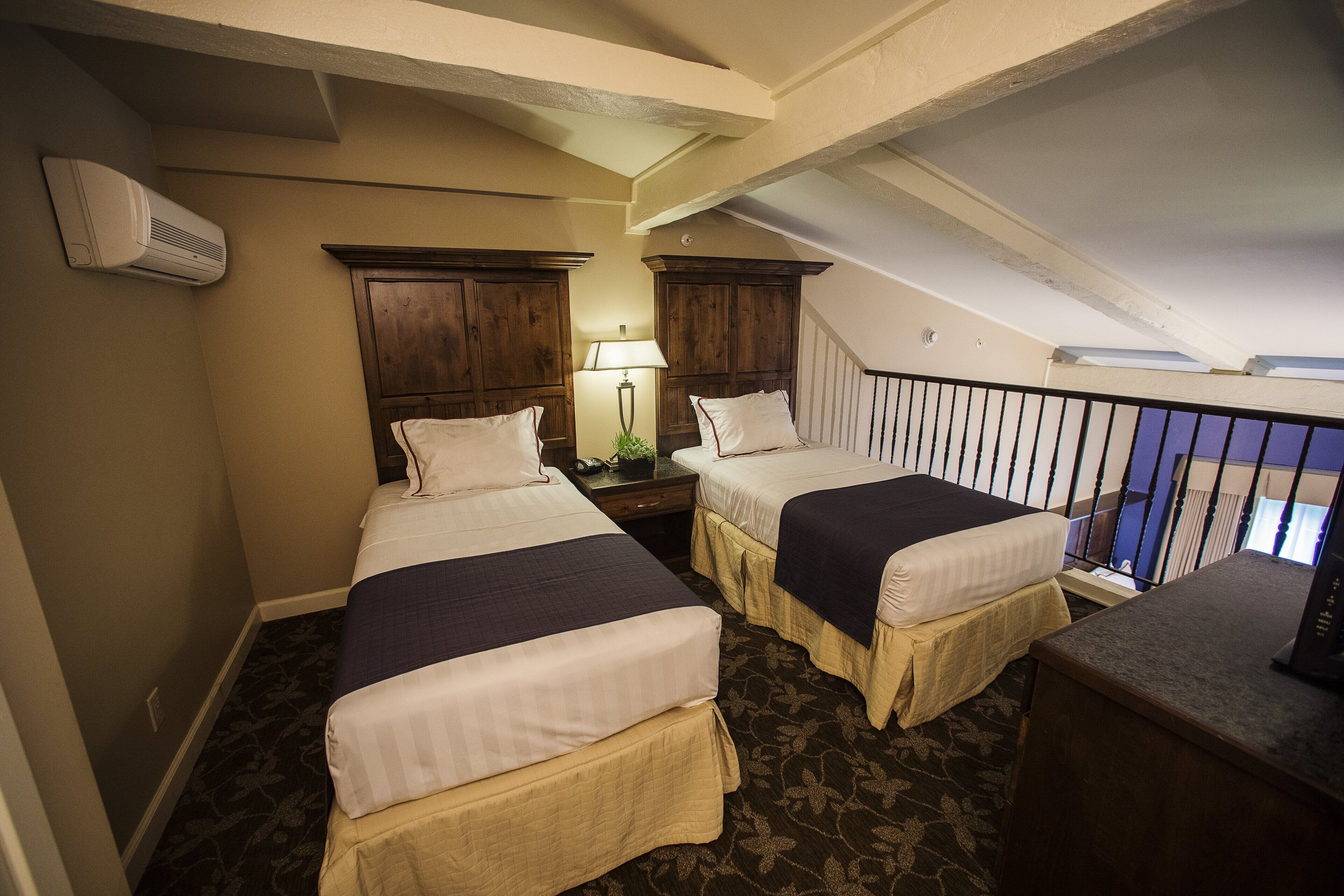 A hotel room with two beds, a desk, a lamp, and a staircase.